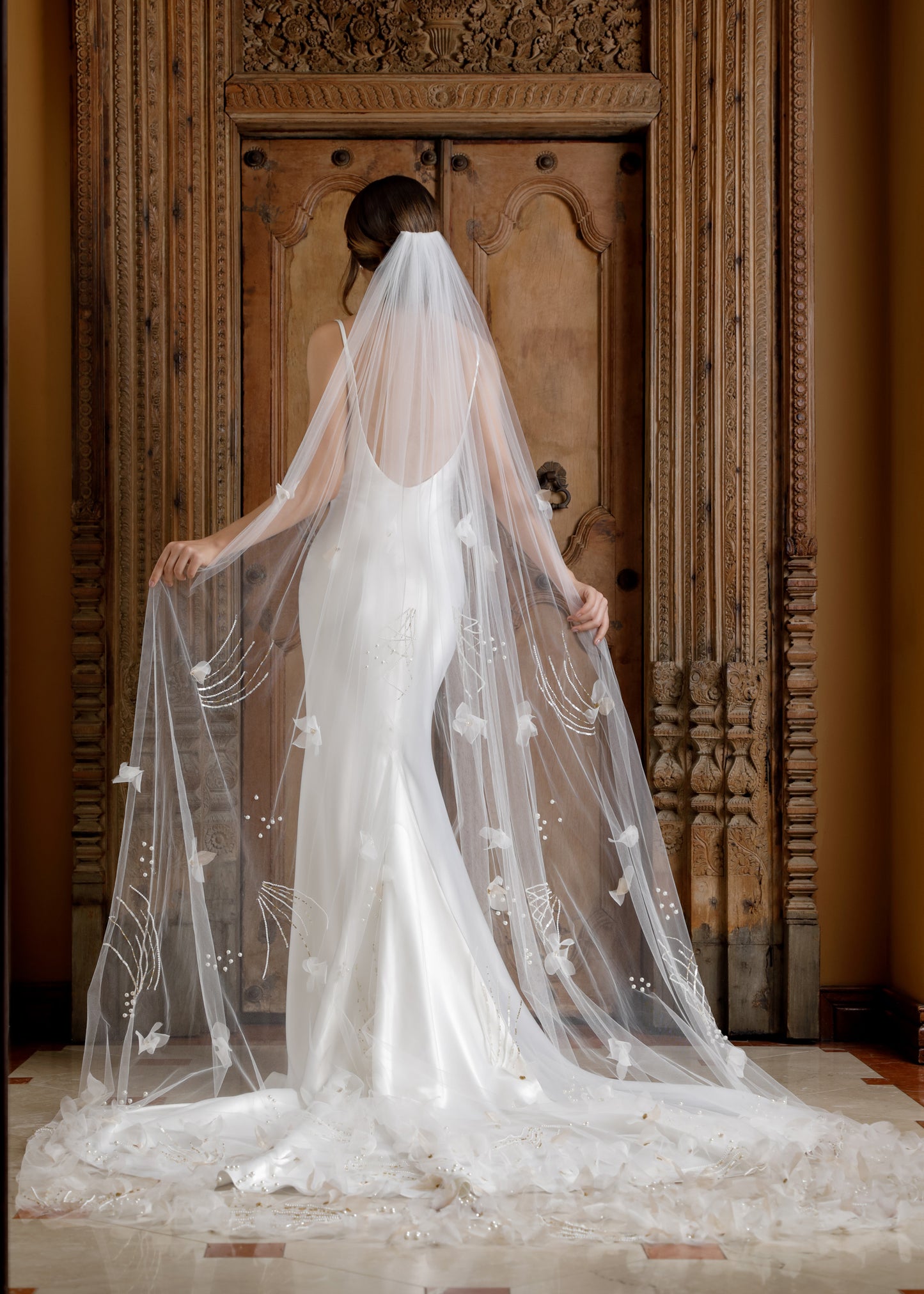 Bride in a white wedding dress with a long veil standing in front of an ornate wooden door.