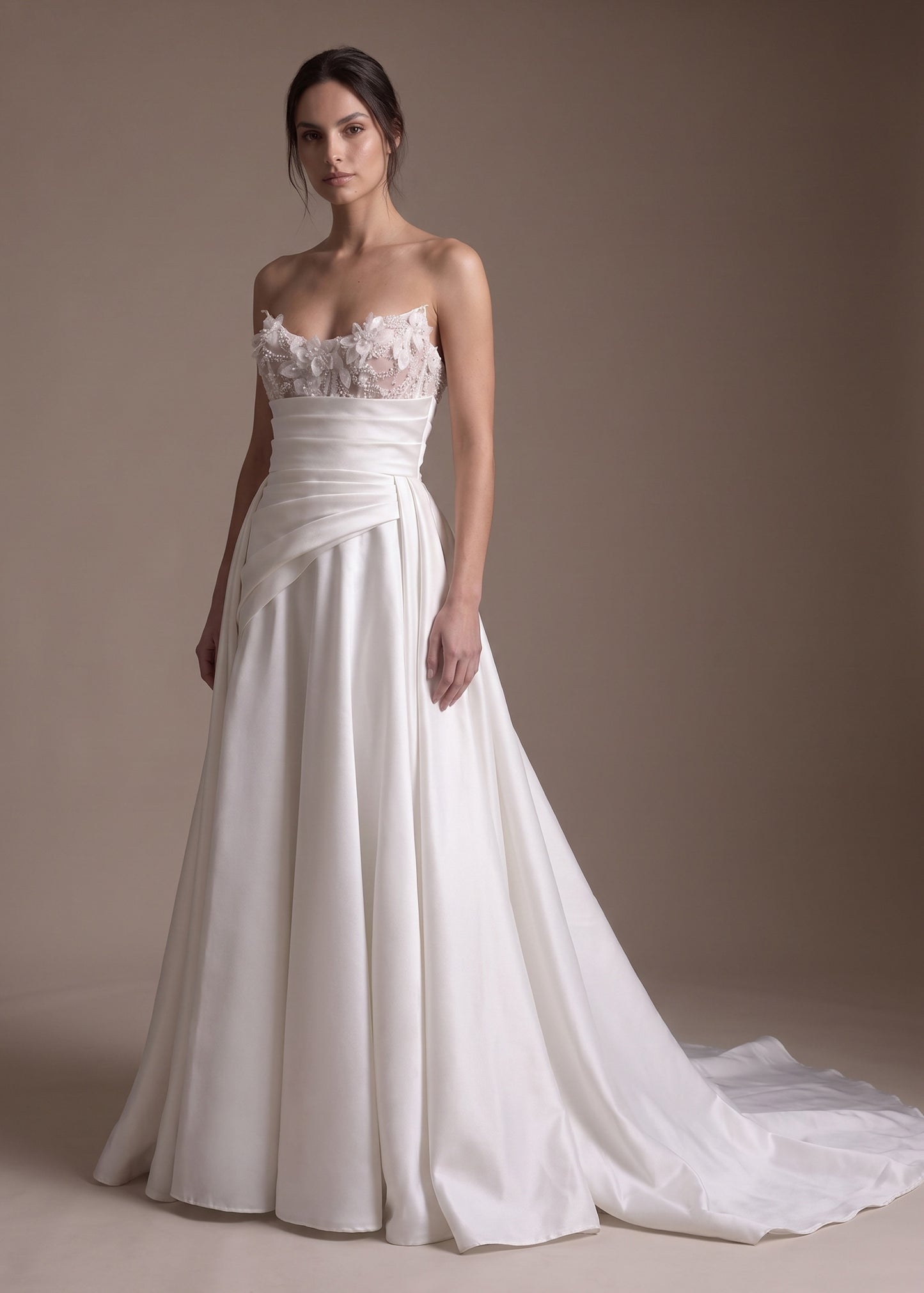 A woman modeling a strapless white wedding gown with a lace sweetheart bodice, draped waist, and full satin skirt with a train, photographed against a plain warm taupe studio background.