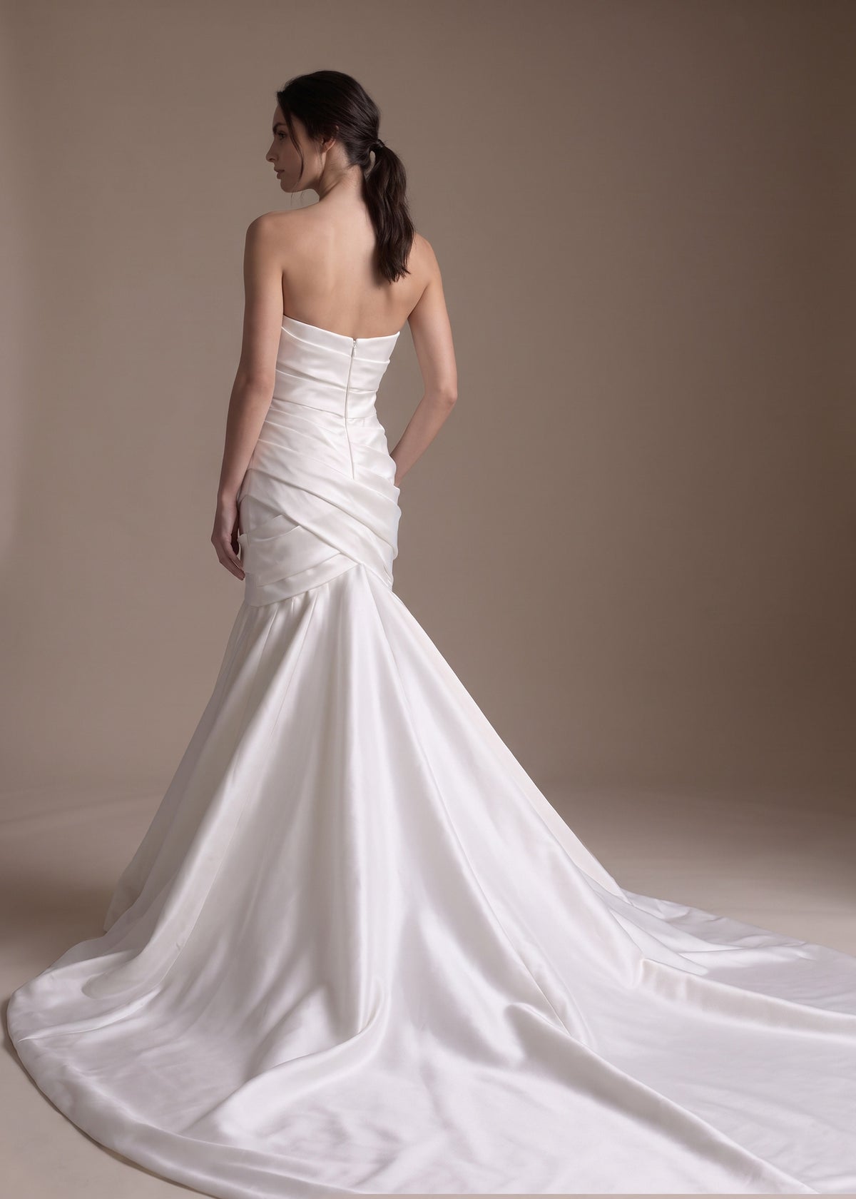 A woman modeling a strapless white satin mermaid-style wedding gown from the back, showing a structured bodice, fitted silhouette, and long flowing train against a warm beige studio background.