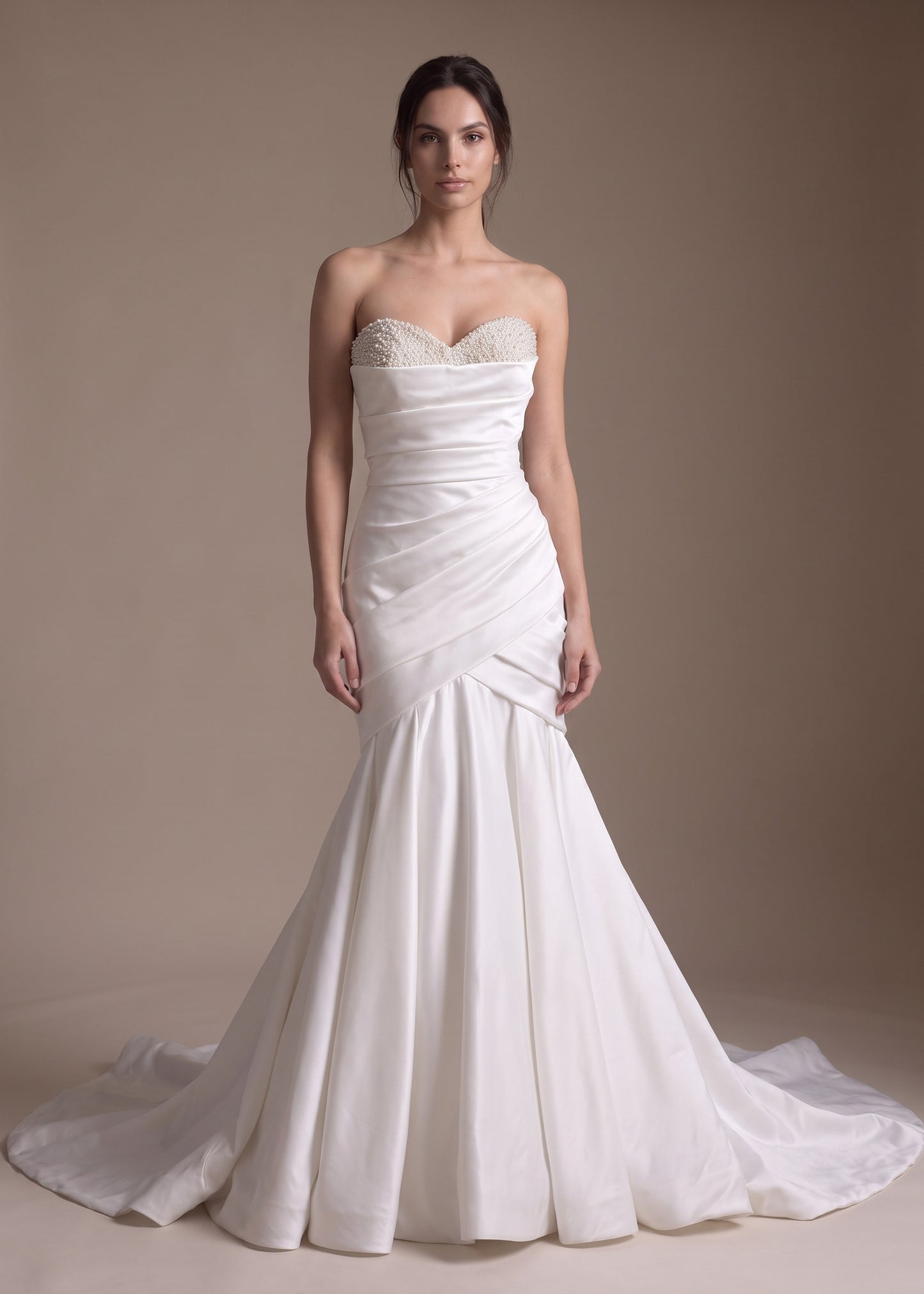 A woman modeling a strapless white mermaid-style wedding gown with a sweetheart neckline, ruched bodice, and slight train against a plain warm beige studio background.