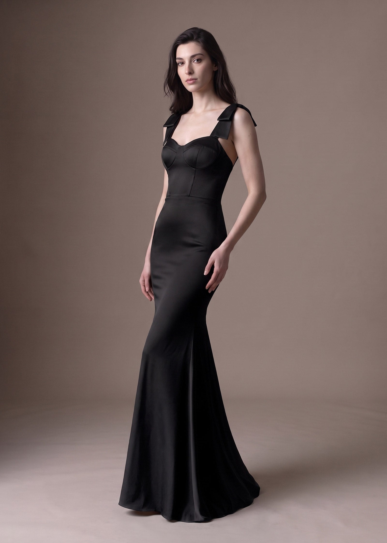 A woman modeling a floor-length black satin gown with structured shoulder straps and a fitted mermaid silhouette, posed against a plain warm taupe studio backdrop.