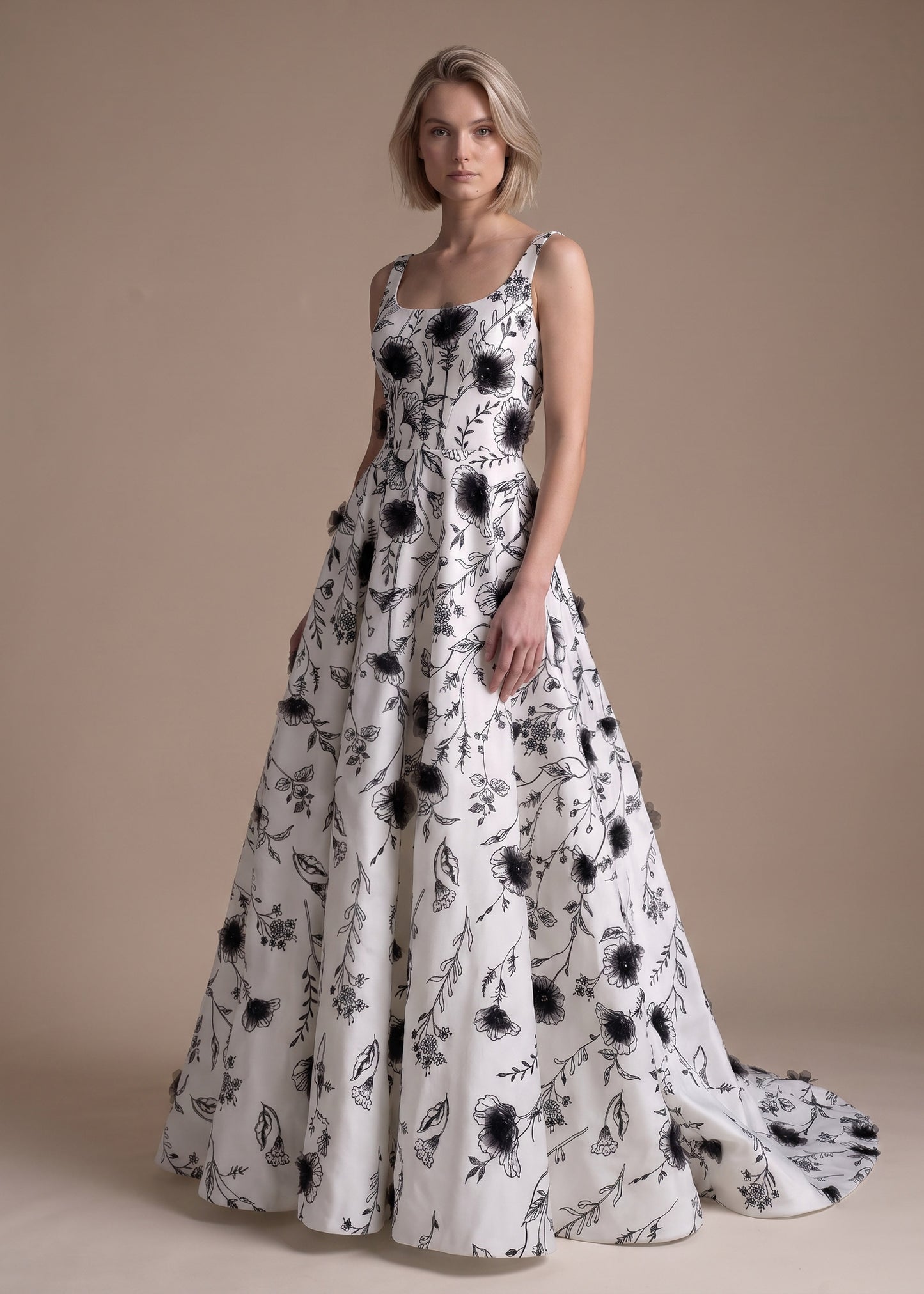 A woman modeling a sleeveless white ball gown with a black floral and leaf pattern, standing against a solid beige studio background.