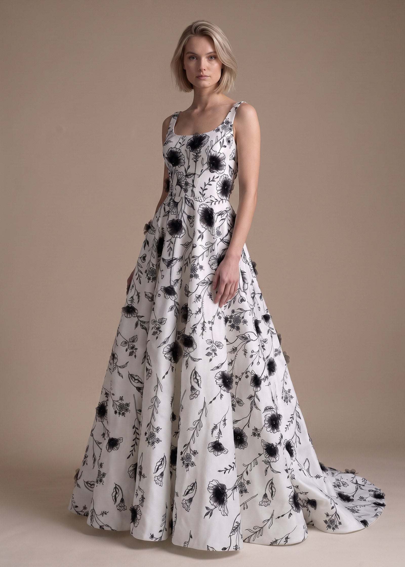 A woman modeling a sleeveless white ball gown with a black floral and leaf pattern, standing against a solid beige studio background.