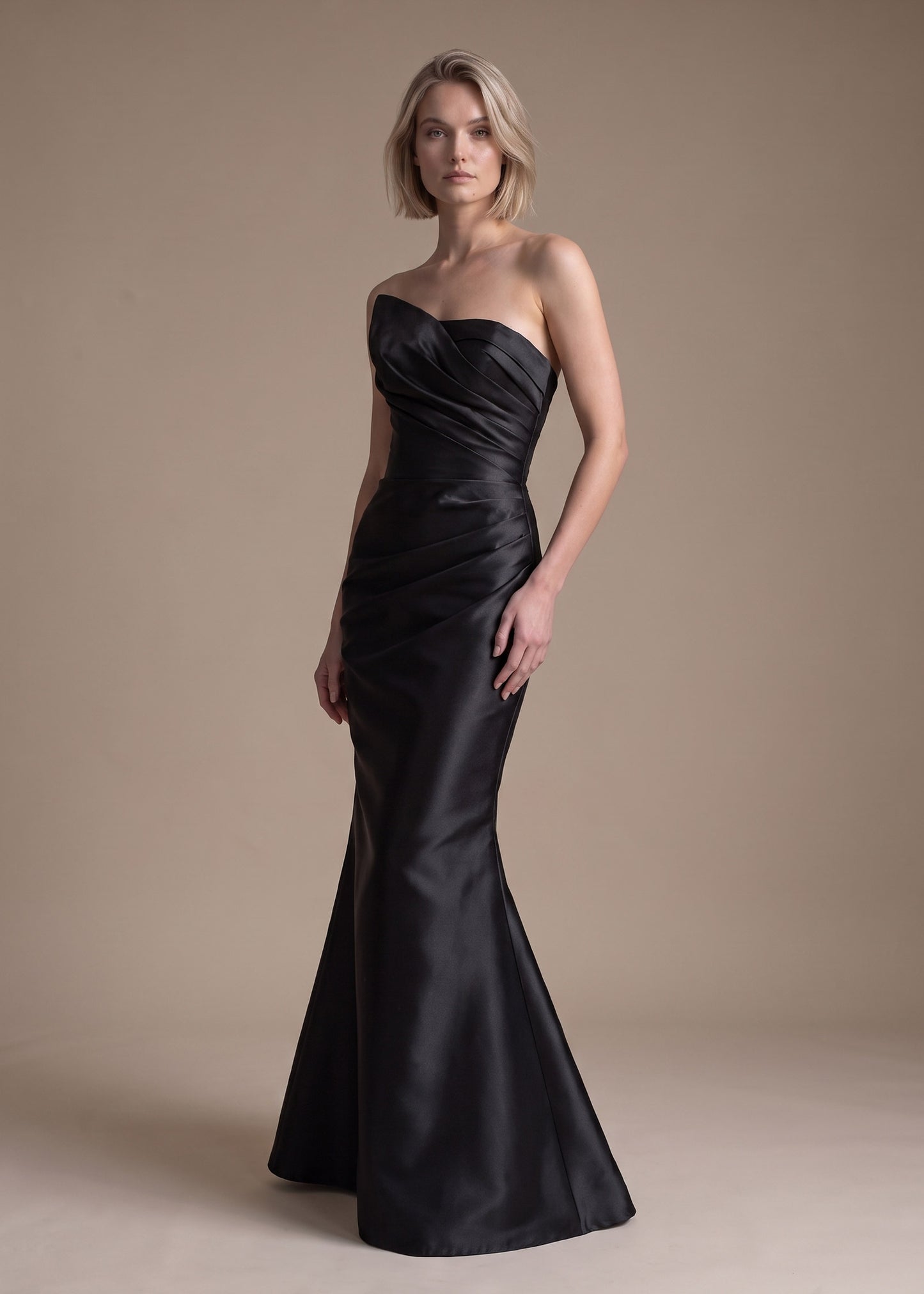 A woman modeling a strapless black satin mermaid gown against a plain beige studio backdrop.