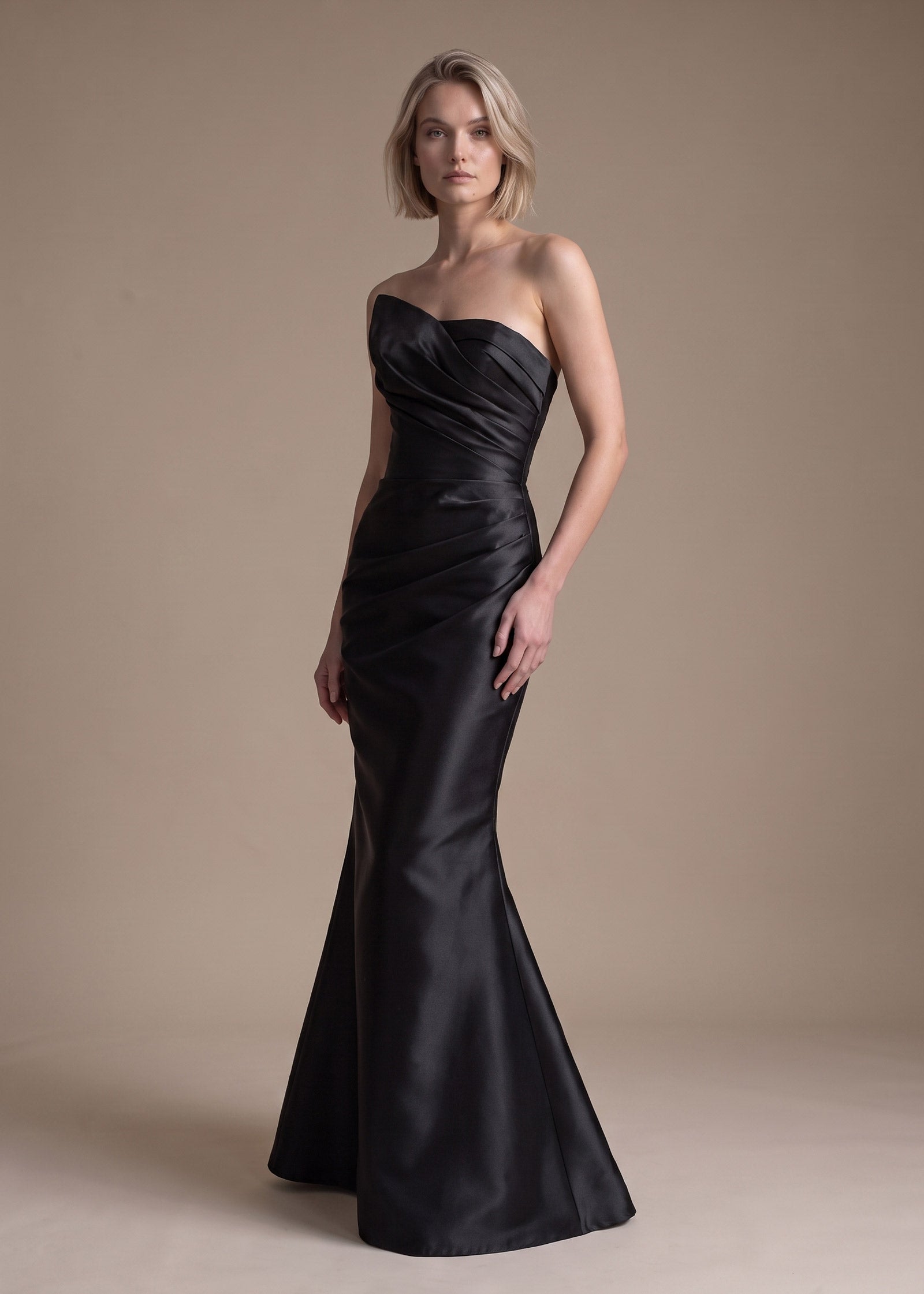 A woman modeling a strapless black satin mermaid gown against a plain beige studio backdrop.