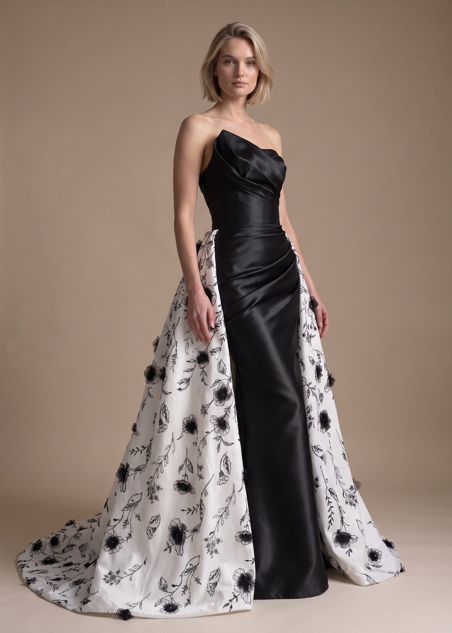 A woman models a strapless formal gown with a black satin bodice and skirt panel, paired with a dramatic white overskirt featuring black floral embroidery and pom-pom trim, against a solid beige studio background.