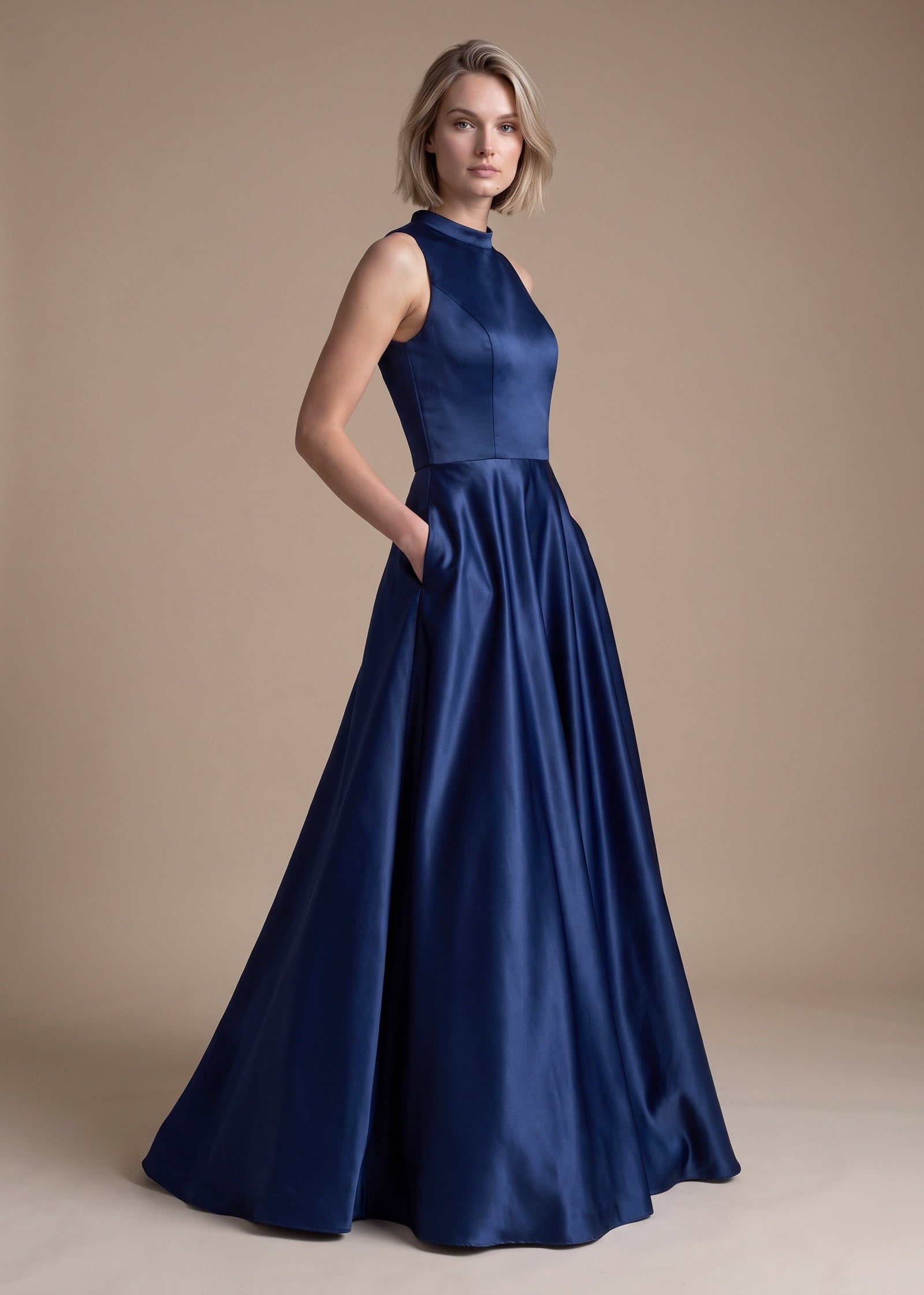 A woman modeling a sleeveless navy blue satin formal gown with a high neckline and a full, flowing skirt against a plain beige studio backdrop.