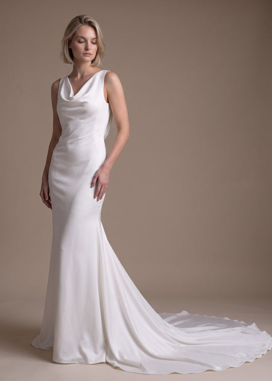 A woman modeling a sleeveless white cowl-neck mermaid gown with a long train against a plain beige studio background.