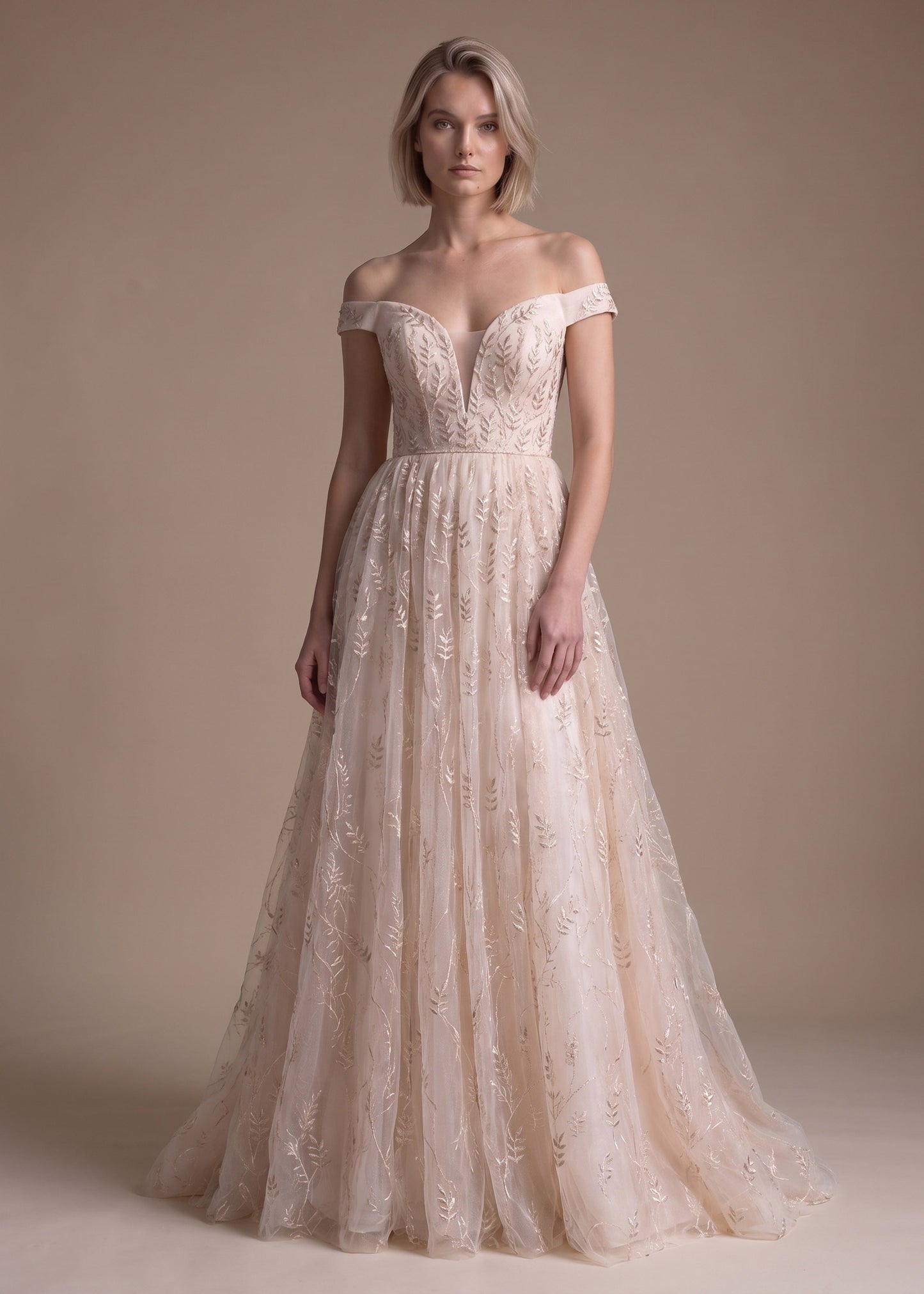 An off-the-shoulder A-line wedding gown in a warm champagne beige, featuring intricate floral lace embroidery on the bodice and cascading tulle skirt, modeled against a solid beige studio backdrop.